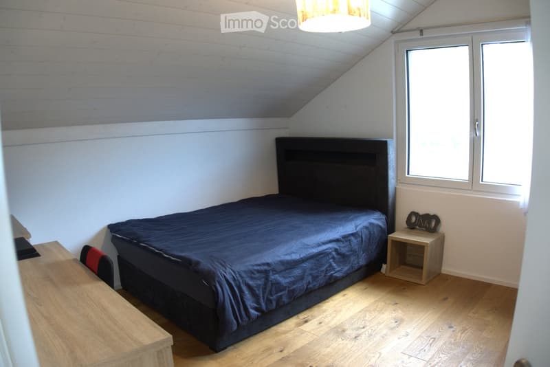 Cozy attic bedroom with wooden floors, slanted ceiling, large windows, a dark-colored bed with a blue duvet, a wooden nightstand, a small table, and a chair.