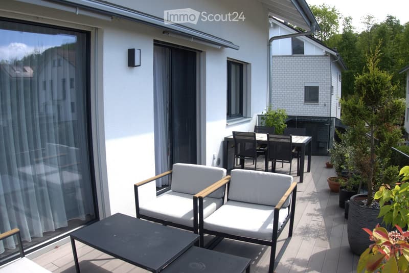 Balcony with white cushioned chairs, a black coffee table, and a dining set with black chairs and a wooden table, surrounded by potted plants.