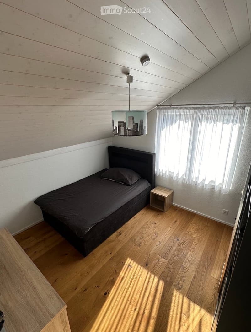 Cozy bedroom with wooden flooring, slanted wooden ceiling, a single bed with dark bedding, a small nightstand, and a hanging lamp with a cityscape design.