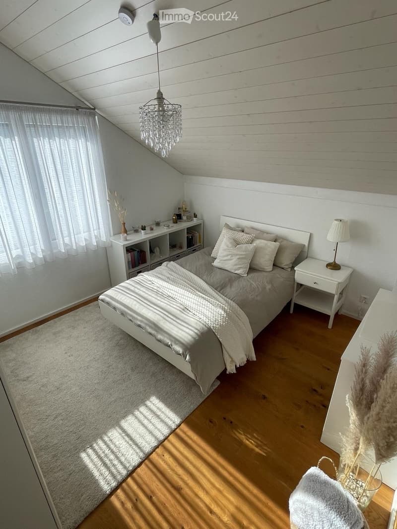 Cozy bedroom with a white bed, nightstand, bookshelf, chandelier, and a soft rug on the wooden floor.