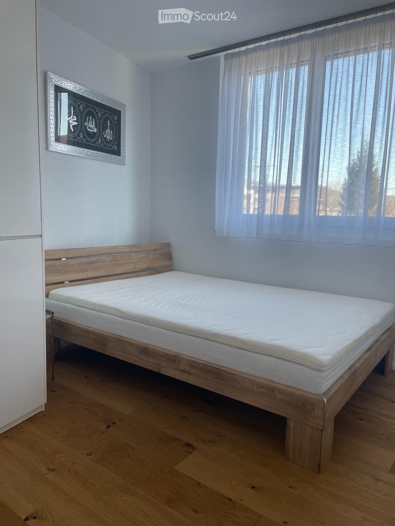 A modern bedroom with a wooden bed frame, a white mattress, a headboard, a large window with sheer curtains, and a decorative mirror on the wall.