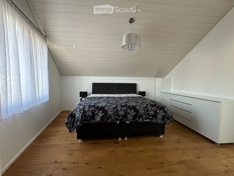 Modern bedroom with wooden flooring, white walls, and a slanted ceiling. Features a black bed with a decorative duvet, a white wardrobe, and a hanging chandelier.