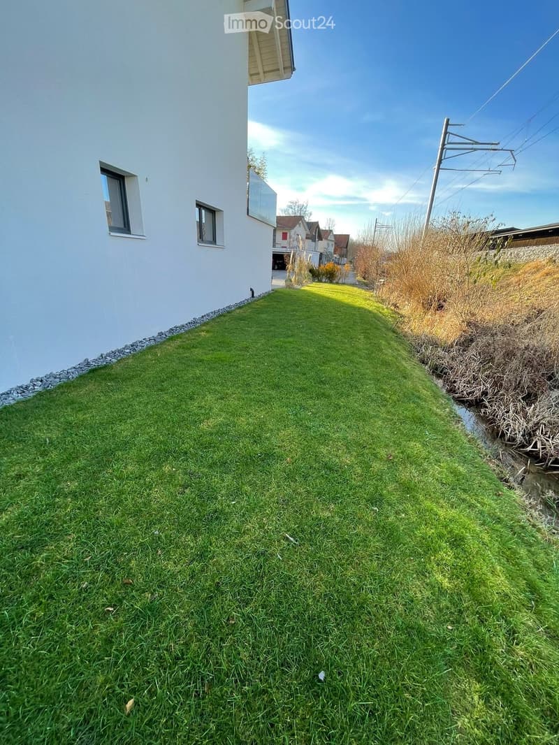 Well-maintained lawn area adjacent to a white building with small windows, featuring a stone border and a small water channel on the right side.