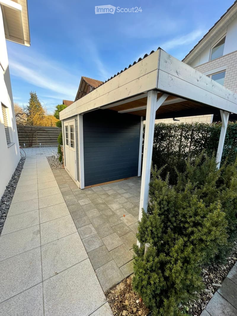 A paved outdoor area with a small garage, a covered patio, and surrounding greenery.