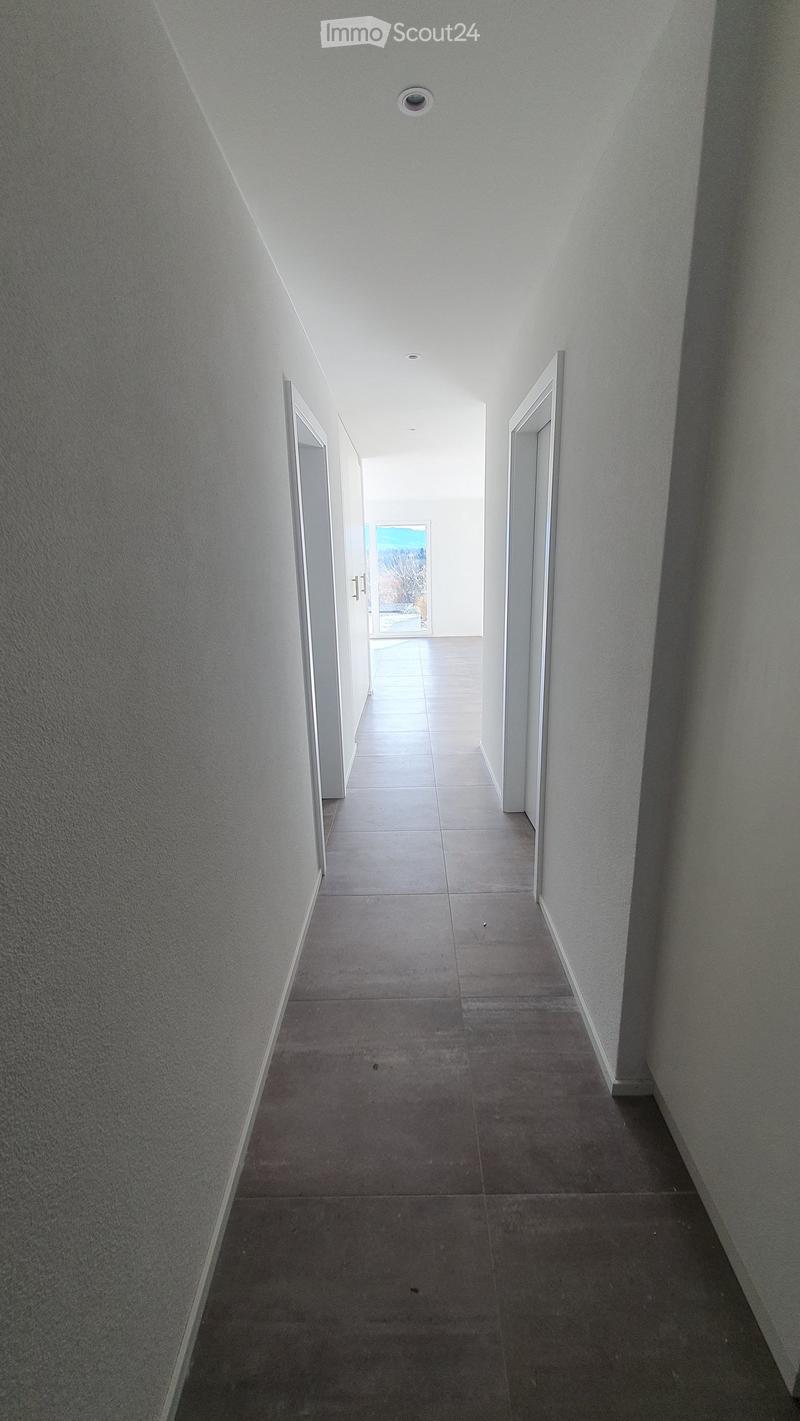 A long, narrow hallway with tiled floors and white walls, featuring several doors and a view of an outdoor area at the end.
