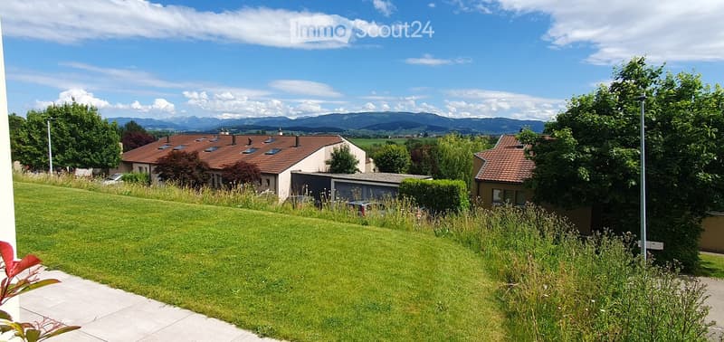 Well-maintained lawn with surrounding trees and shrubs, with houses and a scenic mountain range in the background.