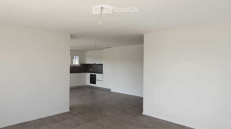 Empty room with white walls, tiled floor, and a small kitchen area with white cabinets and a black countertop.