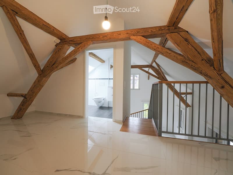 Spacious hallway with exposed wooden beams, marble flooring, and a modern metal railing leading to a staircase. A bathroom is visible through an open doorway.