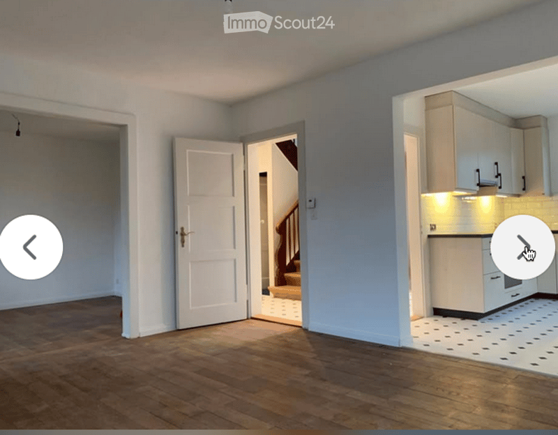 Empty room with wooden flooring and white walls, featuring an open doorway leading to a staircase and a small kitchen area with white cabinets and a countertop.