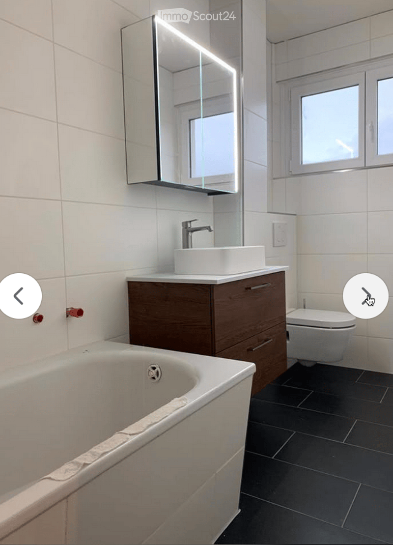 Modern bathroom with white tiles, a bathtub, a sink with a wooden cabinet, a toilet, and a large illuminated mirror above the sink.