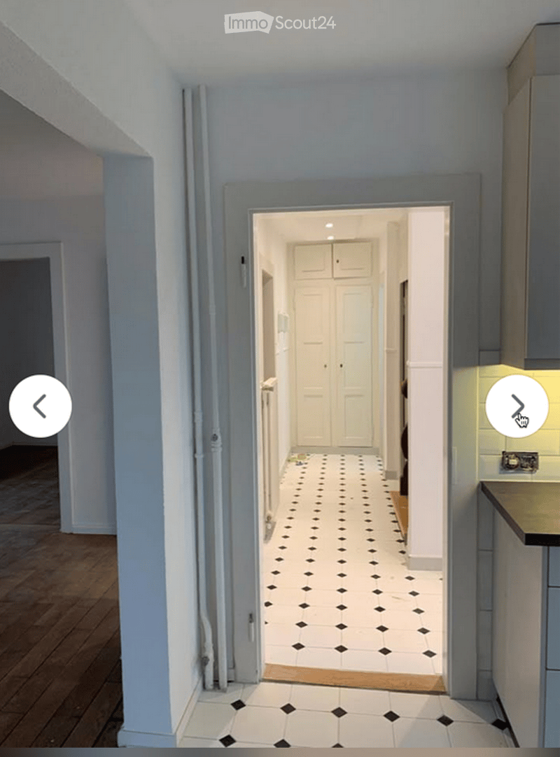 A narrow hallway with a black and white checkered tile floor, leading to a kitchen area with white cabinets and a wooden countertop.