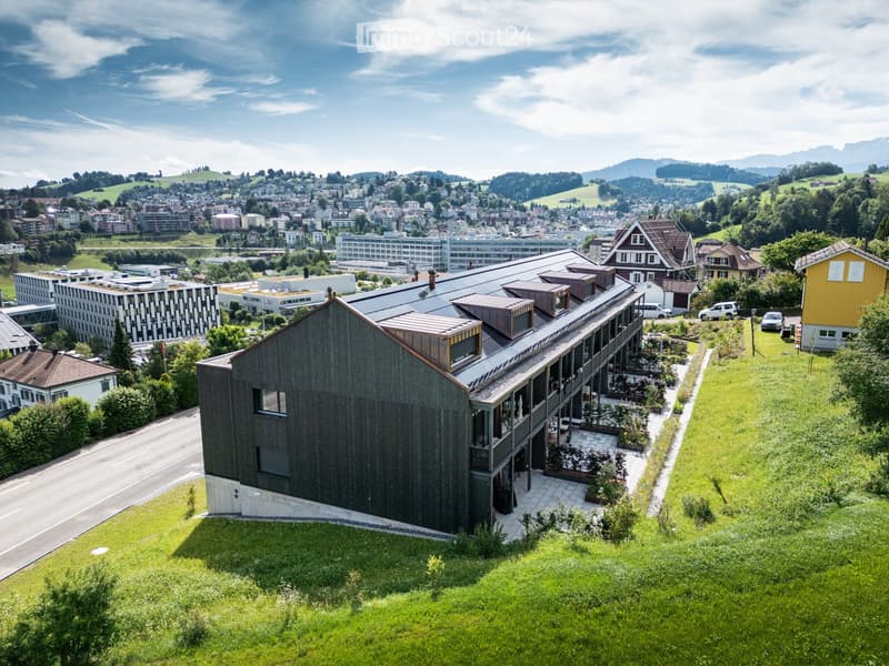 Modern, multi-story building with a sloping roof, black exterior walls, large windows, surrounded by a green lawn with plants and bushes, and visible mountains and a cityscape in the distance.