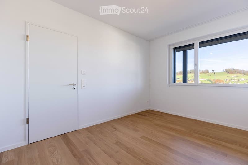 Empty room with white walls, wooden floor, a white door, and a large window with a view of a green field.