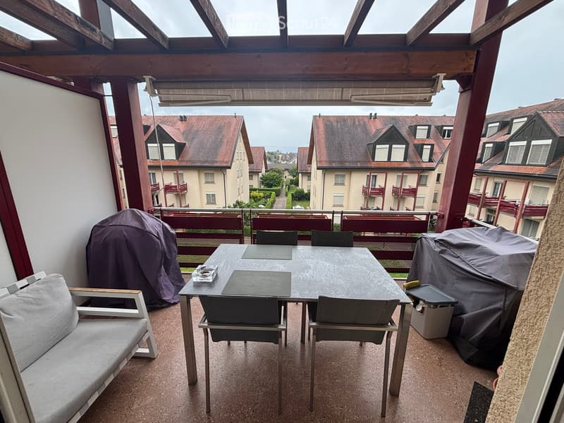 Balcony with a sofa, table, and chairs, overlooking a residential area with apartment buildings.