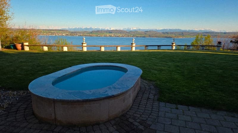 A serene garden with a kidney-shaped swimming pool, surrounded by a well-maintained lawn and a stone balustrade overlooking a lake and distant mountains.