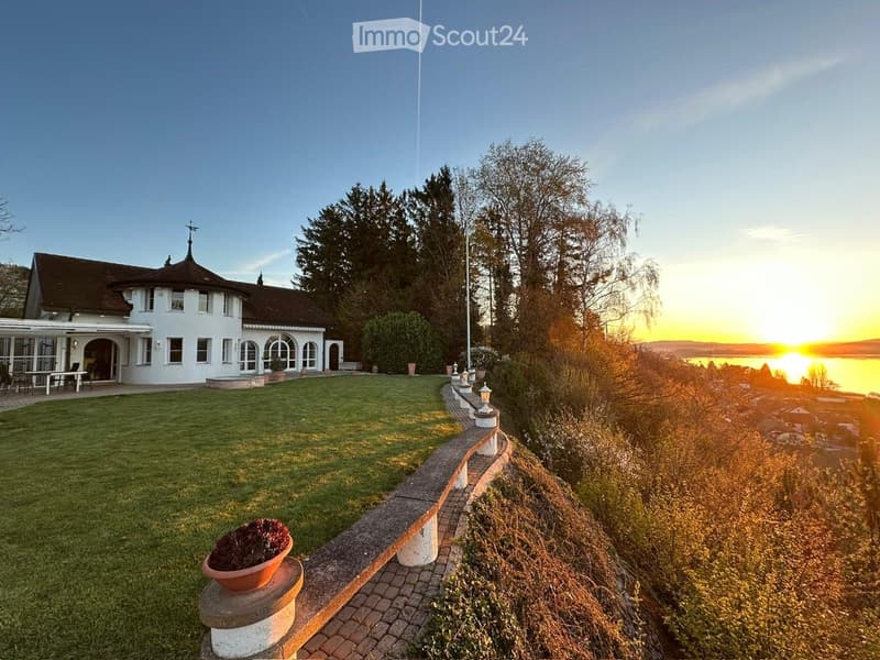 A large, white house with a dome-shaped roof and a balcony overlooking a scenic lake view at sunset.