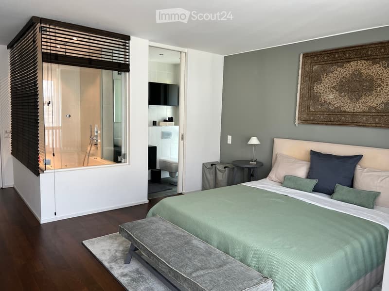 Modern bedroom with a large bed, bench, side table, lamp, and a visible bathroom with a bathtub and toilet.