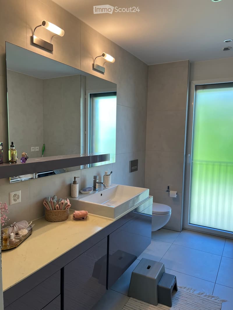 Modern bathroom with a large sink, mirror, and toilet. Features include a wicker basket with towels, a soap dispenser, and two grey stools near the sink.
