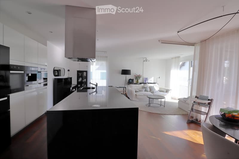 Modern open-plan living and dining area with a white sofa, coffee table, dining table, and chairs. The kitchen features a black island, white cabinets, and stainless steel appliances. Large windows with white curtains allow natural light to fill the space