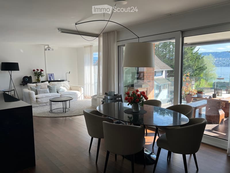 Bright living and dining area with large windows offering a scenic view, featuring a white sofa, coffee table, dining table with chairs, and decorative vases.