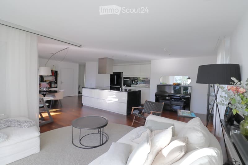 Modern living room with white walls, wooden flooring, and large windows. Features a white sofa with pillows, a round coffee table, a piano, and a kitchen area with white cabinets and black appliances.