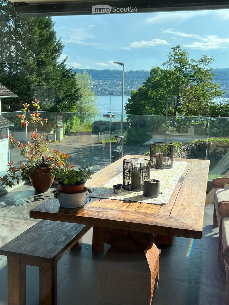 Balcony with wooden table, bench, potted plants, and decorative items, overlooking a scenic view of a lake and city.