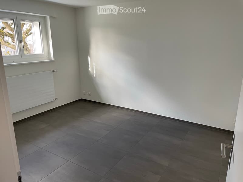 Empty room with grey tiled floor, white walls, a window with white frame, and a radiator below the window.