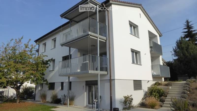 Modern white apartment building with multiple balconies and large windows, surrounded by greenery and a small staircase leading to the entrance.