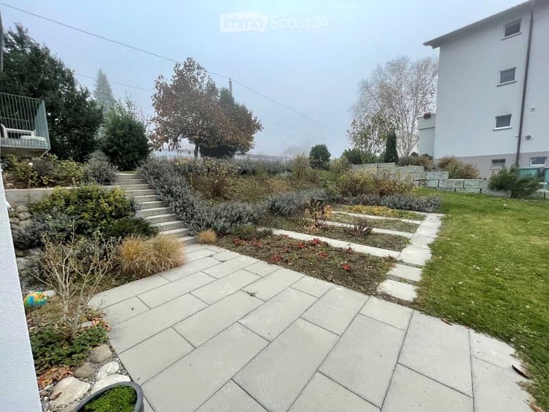 Well-maintained garden with a paved pathway, various shrubs, small trees, and a staircase leading to an upper level. The area includes a small grassy patch and a raised planter with moss.