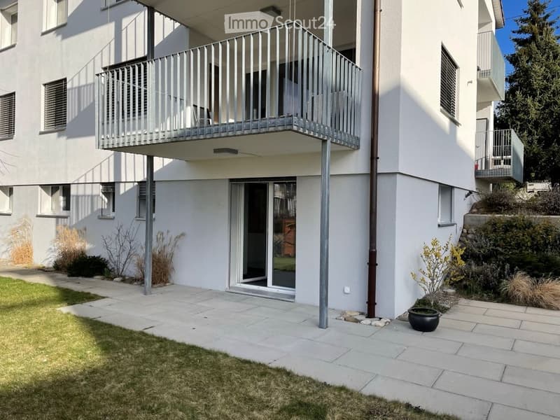 Modern white apartment building with balconies, raised ground floor, and landscaped garden area.