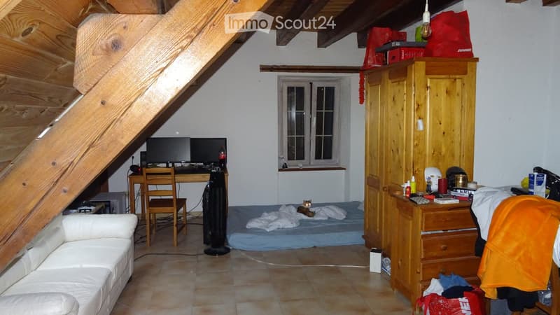 A cozy, multi-functional room with a slanted wooden ceiling, featuring a white sofa, a wooden chair, a desk with a computer, a fan, a small bed with a cat, and a large wooden wardrobe with drawers and various items on top.
