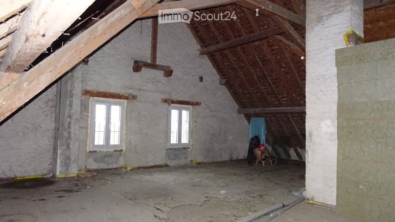 Empty attic space with exposed wooden beams, white plaster walls, and two windows. The floor is unfinished concrete, and there are no furnishings or decorations.
