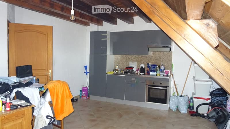 The kitchen features grey cabinets, a stainless steel oven, a coffee maker, and various kitchen items on the counter. There is a wooden chair with an orange blanket draped over it, and a small table with a laptop and other items.