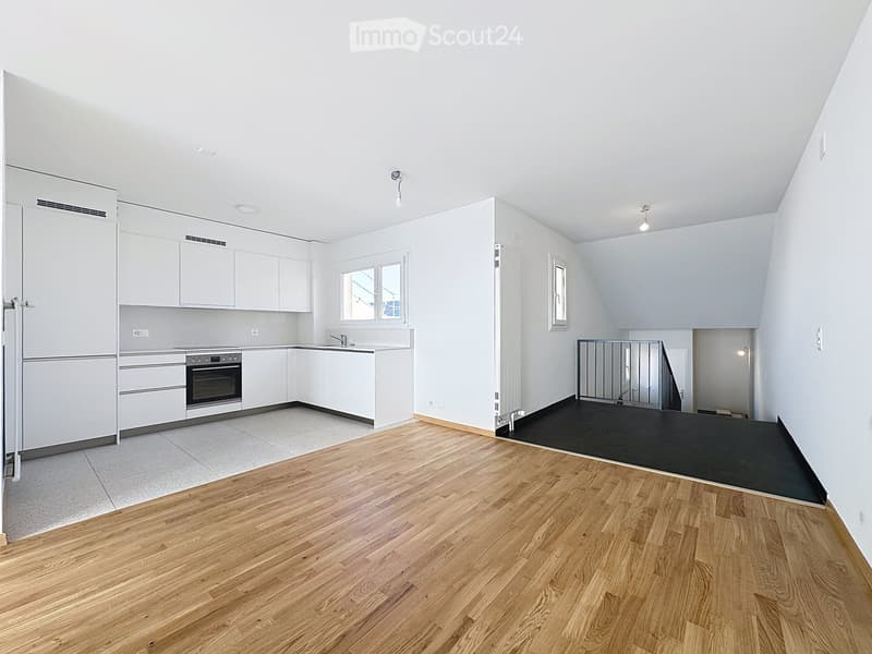 Bright, open room with white walls, wooden flooring, and a small kitchen area featuring white cabinets, a sink, and an oven. The space includes a window and a small black rug in the corner.