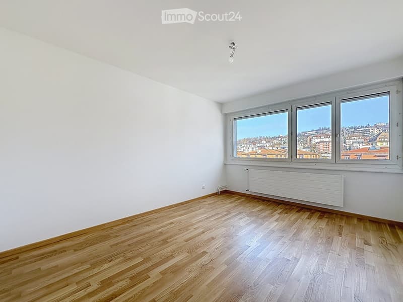 Empty room with wooden flooring, white walls, large windows offering a cityscape view, and a radiator below the windows.