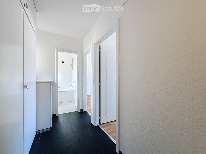A modern hallway with white walls, black tiled floor, and several doors leading to different rooms, including a bathroom with a bathtub and shower.