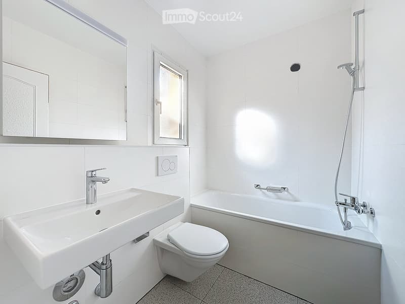 Modern bathroom with white tiles, a sink, toilet, bathtub, and a showerhead. Features a large mirror above the sink and a small window.
