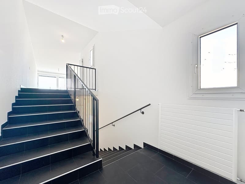 Modern hallway with a staircase featuring dark tiles and a metal railing, white walls, and a large window on the right side.