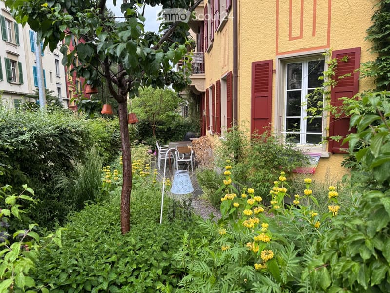 A lush garden area with various plants, flowers, a small table, and chairs, surrounded by colorful buildings with shutters and balconies.