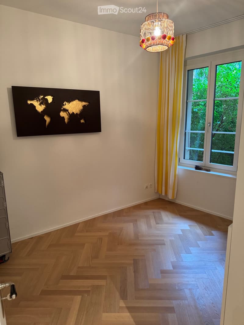 Empty room with wooden herringbone flooring, white walls, large windows with yellow curtains, and a decorative world map painting on one wall.