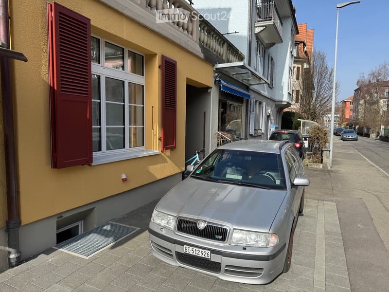A multi-story building with yellow and white facades, red shutters, and a balcony. A silver car is parked in front, and a bicycle is leaning against the building.