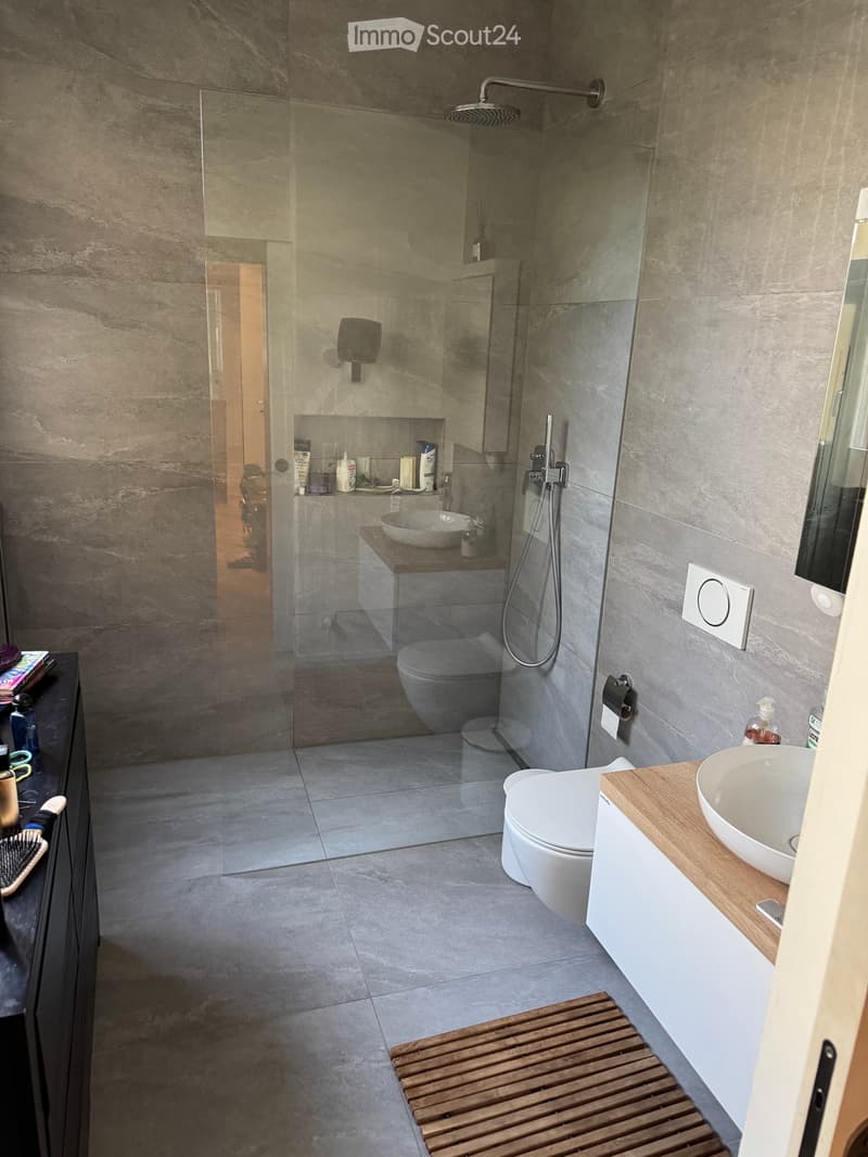 Modern bathroom with grey tiled walls and floor, featuring a glass-enclosed shower, a toilet, a sink with a wooden countertop, and a wooden shelf. A wooden mat is placed in front of the sink.