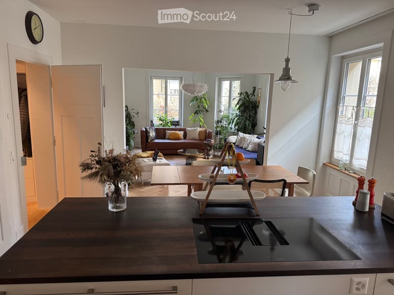 Modern kitchen and living area with a dark wooden island, a sofa, coffee table, and dining area. The space features large windows, a hanging light fixture, and a decorative vase on the island.
