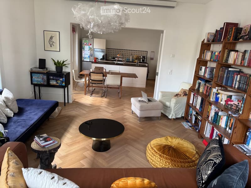 Living room with wooden flooring, a blue sofa, a round coffee table, a gold ottoman, a bookshelf filled with books, a small kitchen area with white cabinets, and a dining table with chairs.
