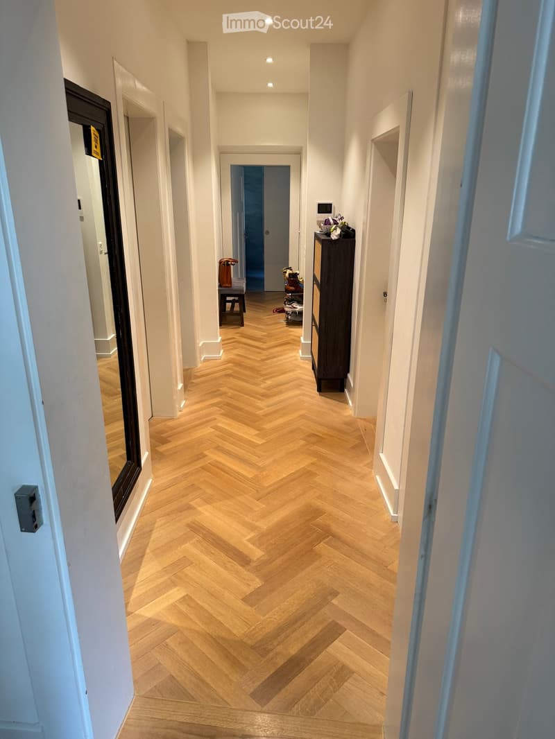 A narrow hallway with herringbone-patterned wooden flooring, white walls, and several doors leading to other rooms.
