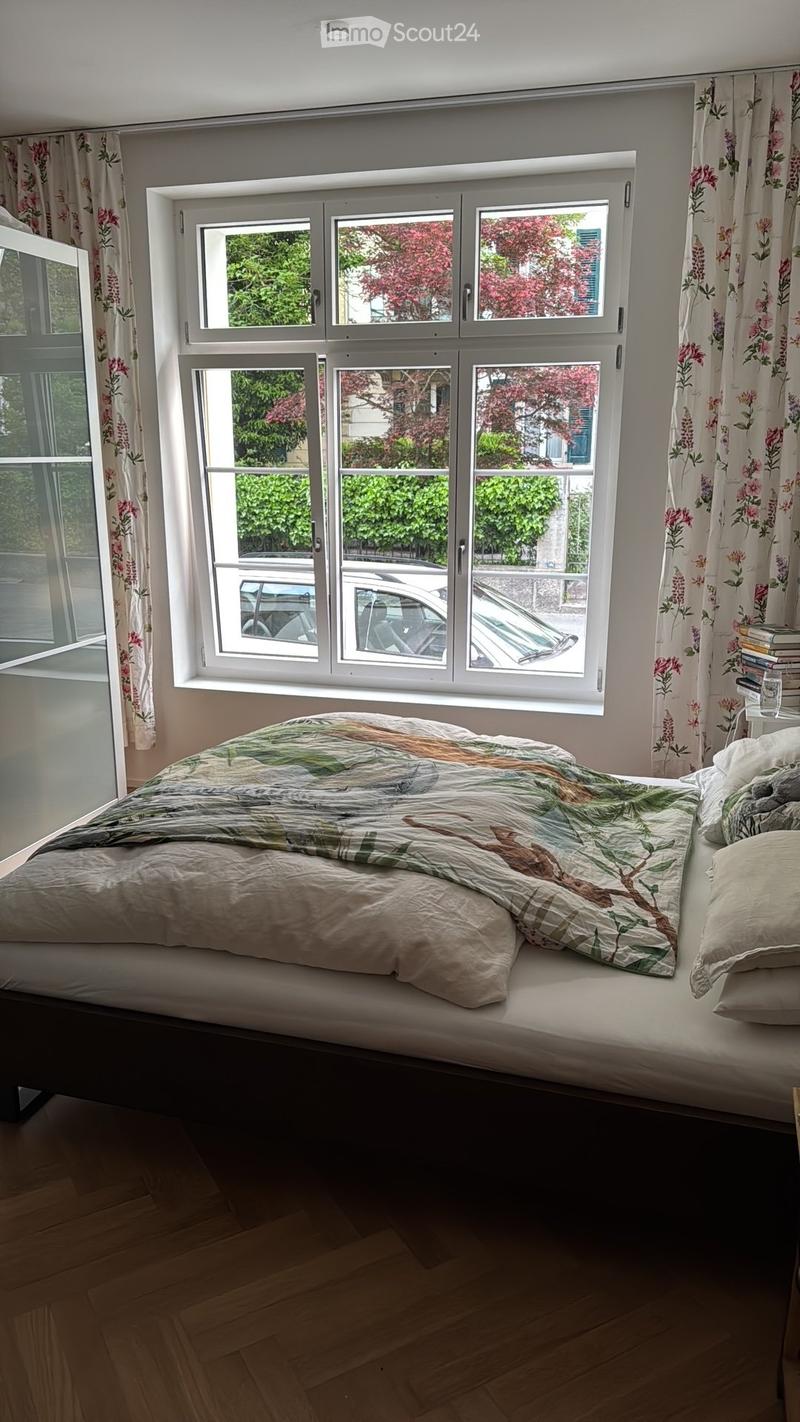 Bright bedroom with large windows, wooden floor, bed with quilt and pillows, nightstand with books and a glass, floral curtains.