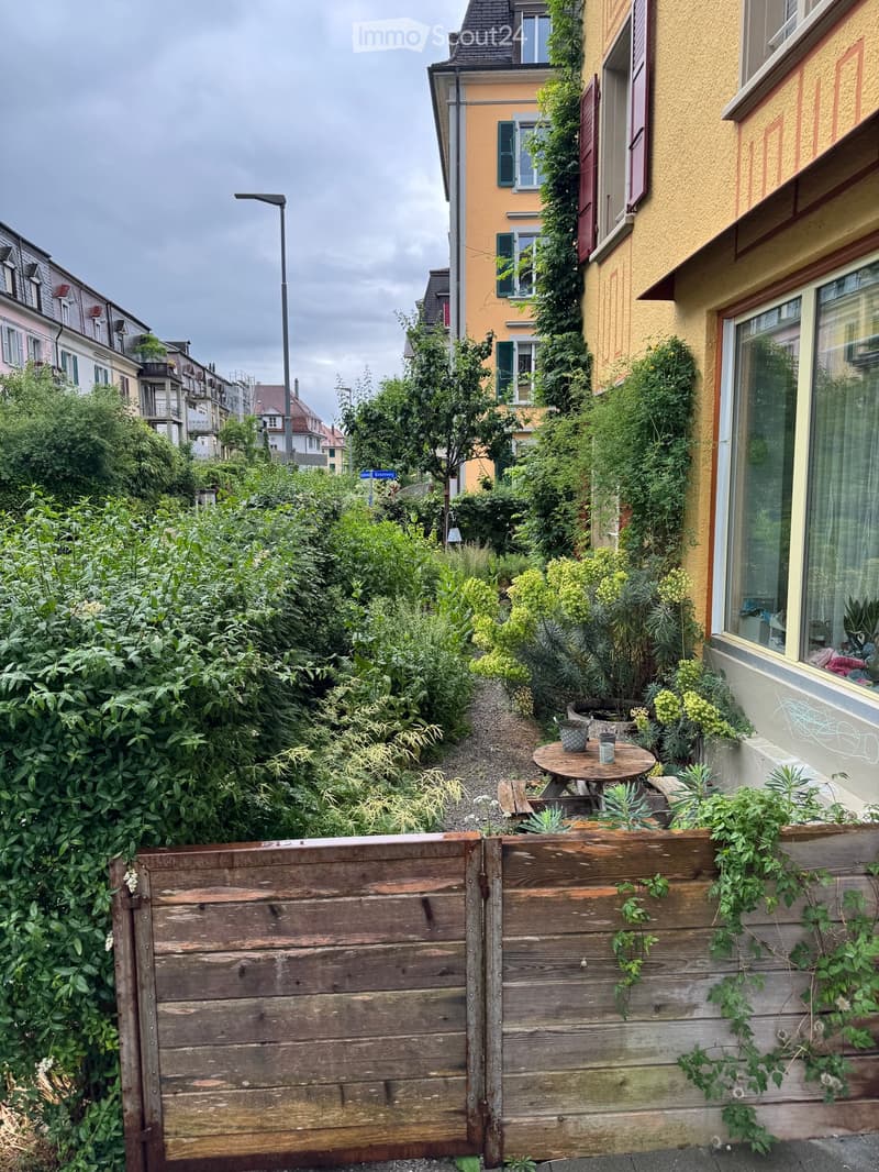 A lush garden area enclosed by wooden planters, with a small round table and chairs, surrounded by greenery and buildings in the background.