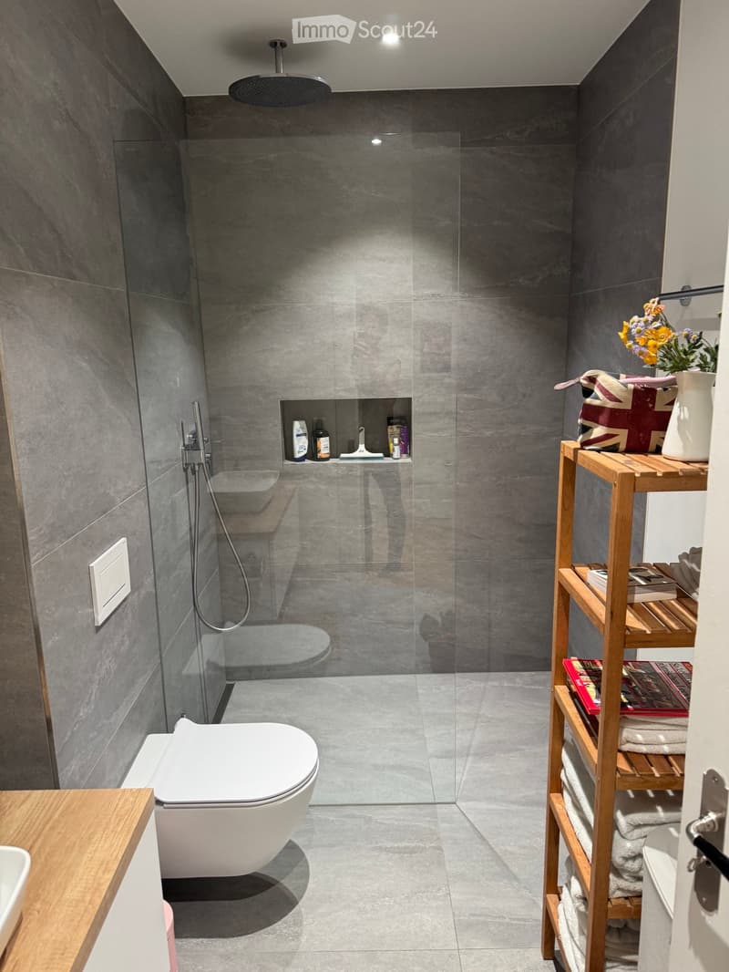 Modern bathroom with grey tiled walls and floor, a white toilet, a glass-enclosed shower area, a wooden shelf holding towels, books, and a vase with flowers, and a small built-in niche for toiletries.