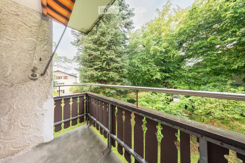 Balcony with metal railing and wooden balusters, overlooking a green garden area with trees and a neighboring house.
