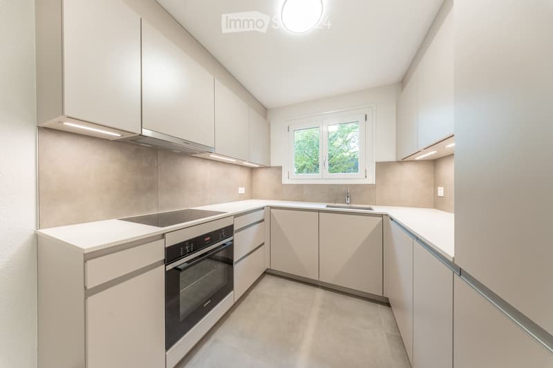 Modern kitchen with white cabinets, stainless steel oven, and a window overlooking greenery.